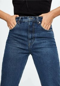 Jeans de denim de tiro alto en azul oscuro con una textura suave, bolsillos frontales y detalles en tono dorado en la cintura y los bolsillos.