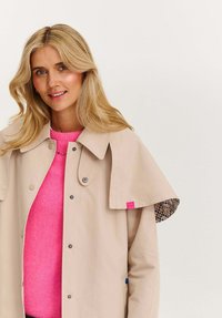 Blondýnka v béžovém trenčkotě, který má přes jasně růžový svetr, stojí proti jednoduchému světlému pozadí a mírně se usmívá.