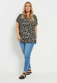Blusa negra con estampado de flores blancas y beige, con mangas cortas y escote abotonado, combinada con jeans ajustados azules y sandalias negras.