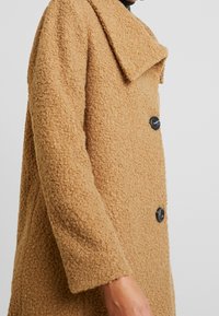 Ingrandimento di una persona che indossa un cappotto di lana texturizzato color sabbia con grandi bottoni neri e un ampio colletto su uno sfondo semplice.