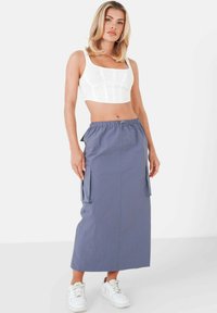 Hellgrauer Cargo-Rock mit einem Kordelzug in der Taille, ausgestattet mit Seitentaschen. Getragen mit einem weißen Cropped-Top und weißen Sneakers. Glatte Stofftextur.