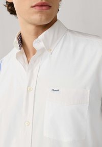 Man draagt een wit overhemd met kraag en knopen, met een zakje waarop 'Façonnable' staat, de knopen van de kraag zijn vastgemaakt en het gestreepte detail aan de binnenkant van de kraag is zichtbaar.