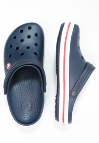 Zoccoli blu navy realizzati in materiale durevole con fori di ventilazione. Presentano strisce bianche e rosse sulla suola e un cinturino flessibile sul tallone.