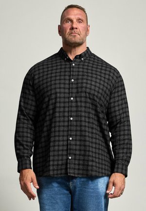 Homme aux cheveux courts et à la barbe, portant une chemise noire à carreaux boutonnée et un jean bleu, debout devant un fond clair uni.