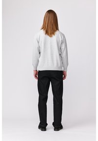 Personne aux longs cheveux bruns portant un pull gris clair, un pantalon noir et des chaussures noires, debout face à un fond blanc uni.