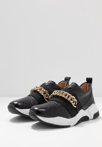Baskets noires en matériau texturé crocodile, avec une sangle élastique et un détail en chaîne dorée. Semelle en caoutchouc blanche et accents texturés noirs.