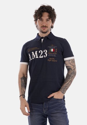 Uomo con capelli ricci che indossa una polo blu scuro con "AM23" e toppe a tema aviazione, e jeans blu, in piedi con una mano in tasca.