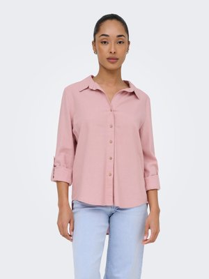 Femme portant une chemise rose à boutons avec les manches retroussées et un jean bleu clair, debout devant un fond blanc uni.