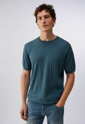 Joven con cabello rizado castaño, vistiendo una camiseta de punto de manga corta verde azulado y jeans azules, de pie con una mano en el bolsillo.