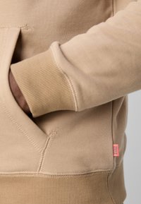 Beige sweatshirt with ribbed cuffs and hem. Features a kangaroo pocket and a small, pink branding tag on the lower side. Smooth texture.