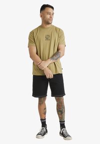 Camiseta de manga corta verde oliva con gráfico floral, acompañada de pantalones cortos negros. El modelo tiene tatuajes y lleva zapatillas negras con cordones blancos.