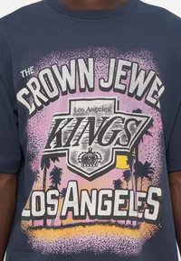 Tricou bleu marin cu un design grafic care afișează "CROWN JEWEL LOS ANGELES KINGS" în alb și roz, împreună cu detalii de palmieri și o coroană.