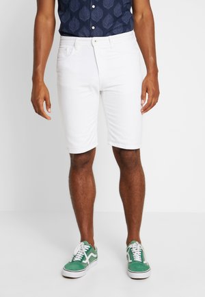 Mann trägt weiße knielange Shorts, grün-weiße Turnschuhe und ein dunkelblaues Kurzarmhemd mit Blattmuster und steht auf weißem Boden.