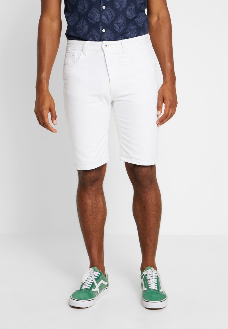 Homme portant un short blanc mi-long, des baskets vertes et blanches, et une chemise bleu foncé à manches courtes avec un motif de feuilles, debout sur un sol blanc.