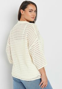 Vit löst stickad tröja med trekvartsärmar, som har en avslappnad passform och en andningsbar textur, ihop med ljusblå jeans.