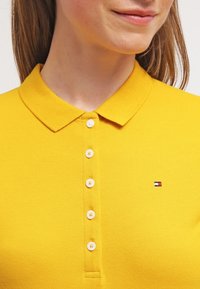 Gul polo-shirt i ett strukturerat tyg, med krage och sex vita knappar, samt en liten, flerfärgad logotyp på vänster bröst.