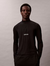 Črn dolg rokav vrh z ovratnikom na pol zadrgo in malim belim logotipom "CALVIN KLEIN" na prsih, izdelan iz gladkega, lahkega materiala.