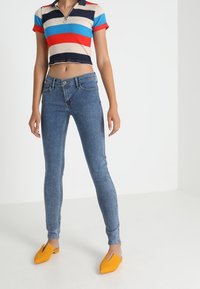 Mujer vistiendo jeans ajustados azules, una camiseta polo recortada a rayas coloridas y unos zapatos planos amarillos mostaza de punta, de pie sobre un suelo blanco.