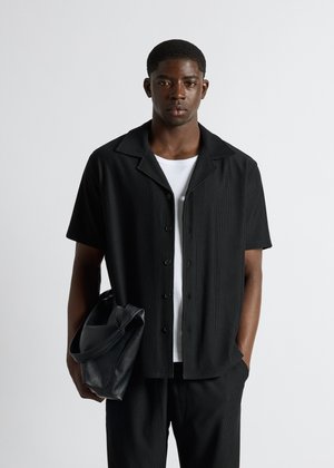 Mann trägt schwarzes kurzärmeliges Hemd mit Knöpfen über weißem T-Shirt, schwarze Hose, hält schwarze Ledertasche vor hellem einfarbigem Hintergrund.