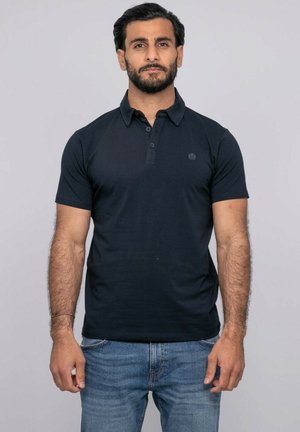 Homme aux cheveux foncés et à la barbe, portant un polo noir à manches courtes et un jean bleu, debout devant un fond gris uni.