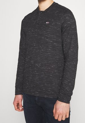 Homme portant un t-shirt henley à manches longues gris foncé avec trois boutons et un petit logo sur la poitrine, accompagné d'un jean bleu foncé.