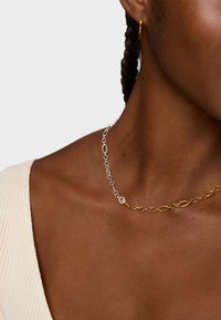 Srebrno-złota bransoletka z ogniwami w kształcie owalnym, podzielona na naprzemiennie ułożone elementy. Cienki, delikatny design opiera się na obojczyku, uzupełniony prostym zapięciem.