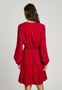 Robe rouge en tissu texturé, avec des manches longues, une taille cintrée et un ourlet volants, vue de dos.