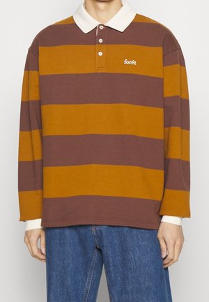 Mand iført brun og sennepsfarvet rugbytrøje med brede striber, hvidekrave og "forêt"-logo, kombineret med blå jeans.