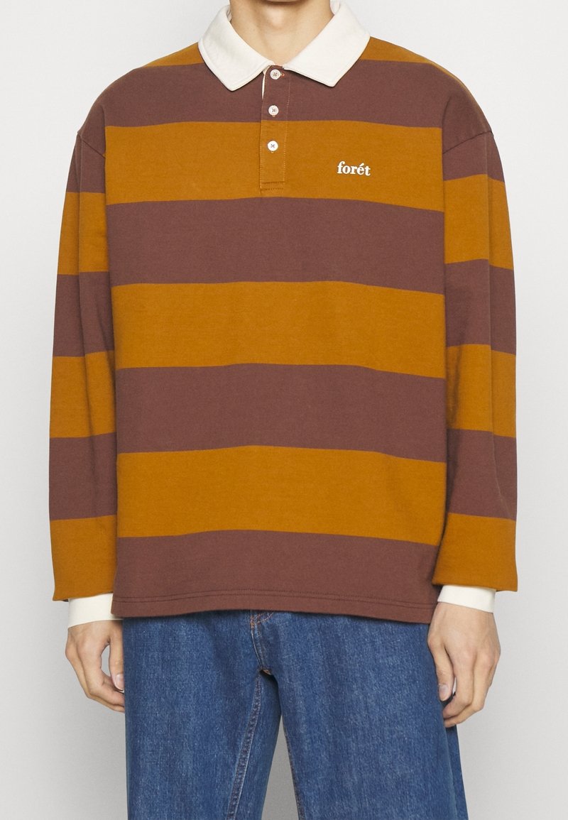 Homme portant un maillot de rugby à manches longues à larges rayures marron et moutarde avec un col blanc et un logo « forêt », assorti à un jean bleu.