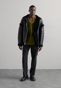 Black leather jacket with shearling collar, green patterned knit sweater, and slim-fit black trousers with white piping. Black shoes.