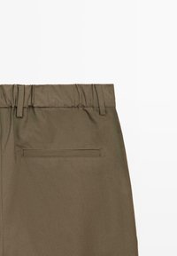 Primo piano di pantaloni verde oliva che mostrano una vita elastica con passanti per cintura e una tasca posteriore a filetto singolo su uno sfondo neutro.