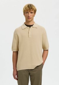 Homme blond portant un polo en maille beige à manches courtes sur un sous-shirt foncé avec un pantalon vert olive, debout devant un fond blanc uni.