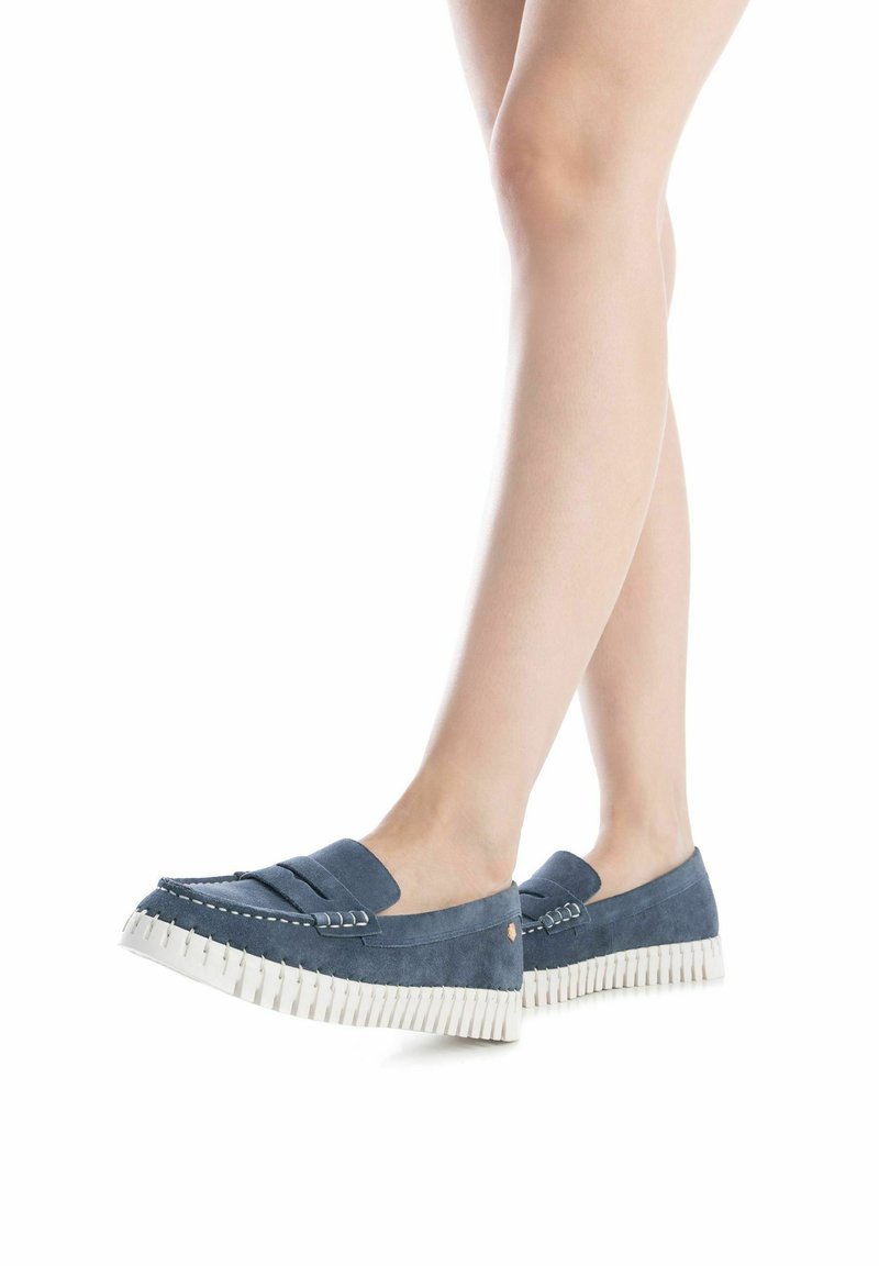 Mocasines de ante azul con una suela blanca dentada, que presentan un acento cosido y un detalle de sujetador de moneda, situados sobre un fondo neutro.