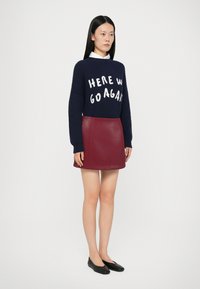 Maglione blu navy a maglia con slogan bianco "ECCOCI ANCORA," abbinato a una mini gonna in pelle bordeaux. Ballerine nere completano il look.