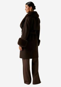 Cappotto di lana marrone con ampio colletto e polsini in pelliccia, vita cinturata e taglio fino al ginocchio. Tessuto liscio, disegnato con una silhouette moderna.