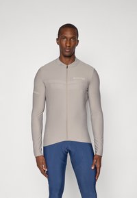 Koszulka rowerowa z długim rękawem w jasnoszarym kolorze z zamkiem błyskawicznym z przodu, o slim fit i minimalistycznym logo. W zestawie z niebieskimi szortami rowerowymi.