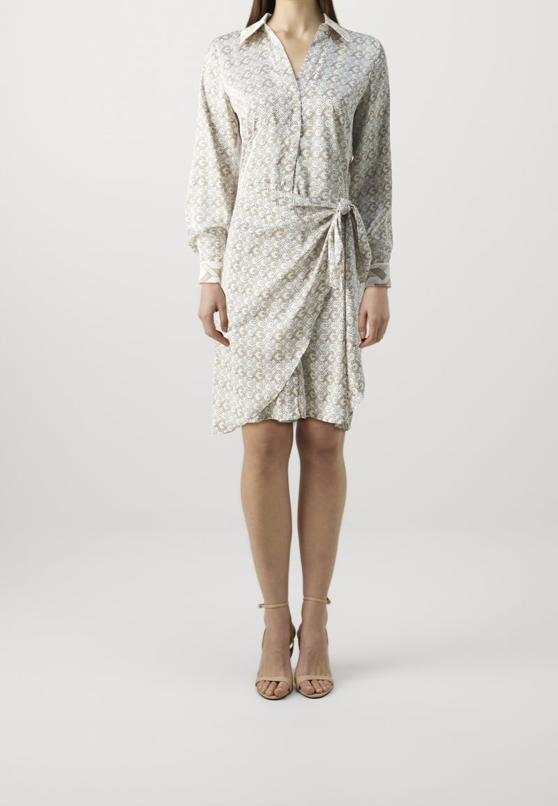 Femme portant une robe à manches longues boutonnée avec un motif, un détail de nœud sur le côté et des sandales à talons beiges, debout devant un fond uni.