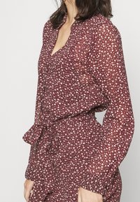 Mörkröd blommig klänning med ett lätt, genomskinligt tyg. Den har långa ärmar, en v-ringning och en knytning i midjan. Små blommönster över hela klänningen.