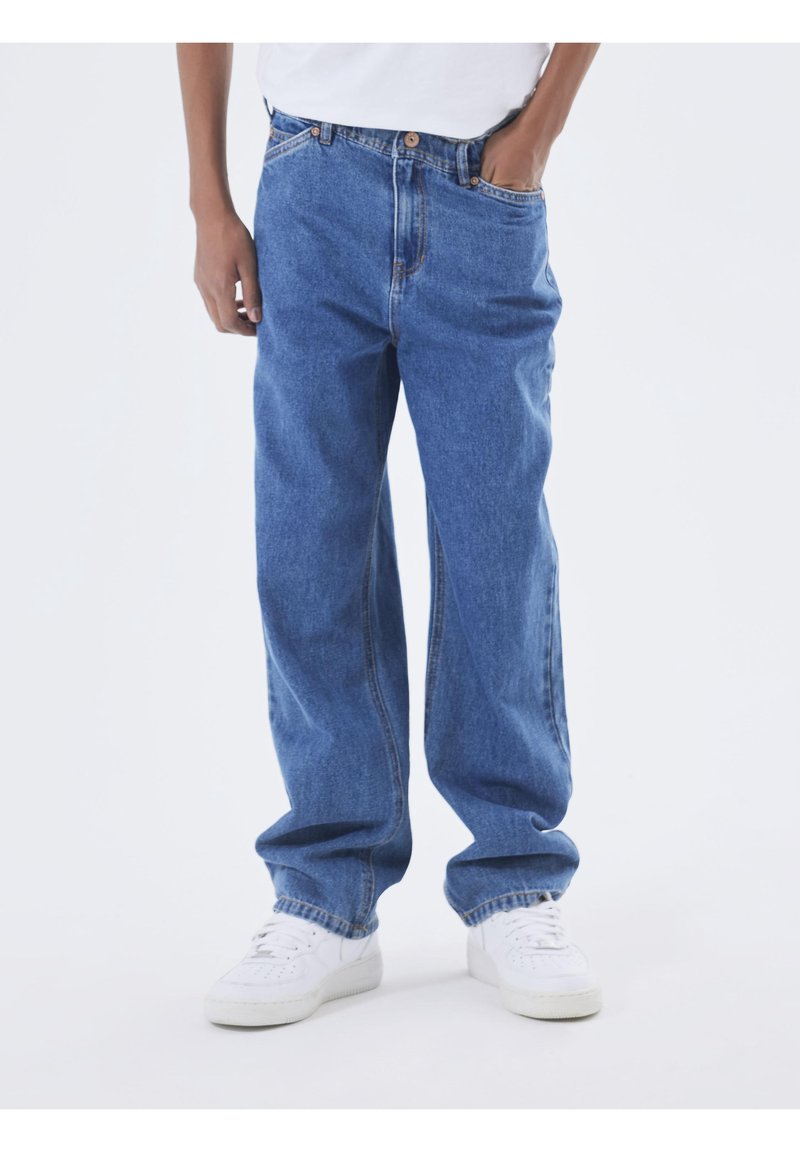 Personne portant un jean bleu ample et des baskets blanches, debout avec une main dans la poche sur un fond clair uni.