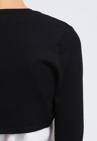 Nahaufnahme einer Person, die einen schwarzen, kurzen Pullover über einem weißen, strukturierten Kleidungsstück trägt, wobei Rücken- und Schulterdetails zu sehen sind.