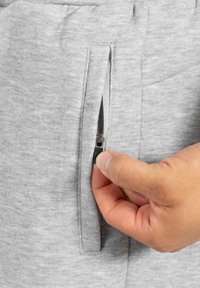 Pantalones de chándal grises con un acabado texturizado, con un bolsillo lateral con cremallera que está siendo abierto por una mano.