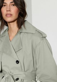 Trench coat grigio chiaro con ampio colletto, doppi bottoni e vita con cintura, caratterizzato da una texture liscia e da un leggero effetto a sbuffo sulle maniche.