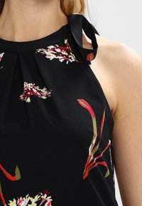 Musta toppi, jossa on punainen, vihreä ja valkoinen kukkakuosi. Kaksiosainen kaula-aukko, jossa on solmittava kaulaosa ja poimutuksia kaula-aukossa tekstuurin luomiseksi.