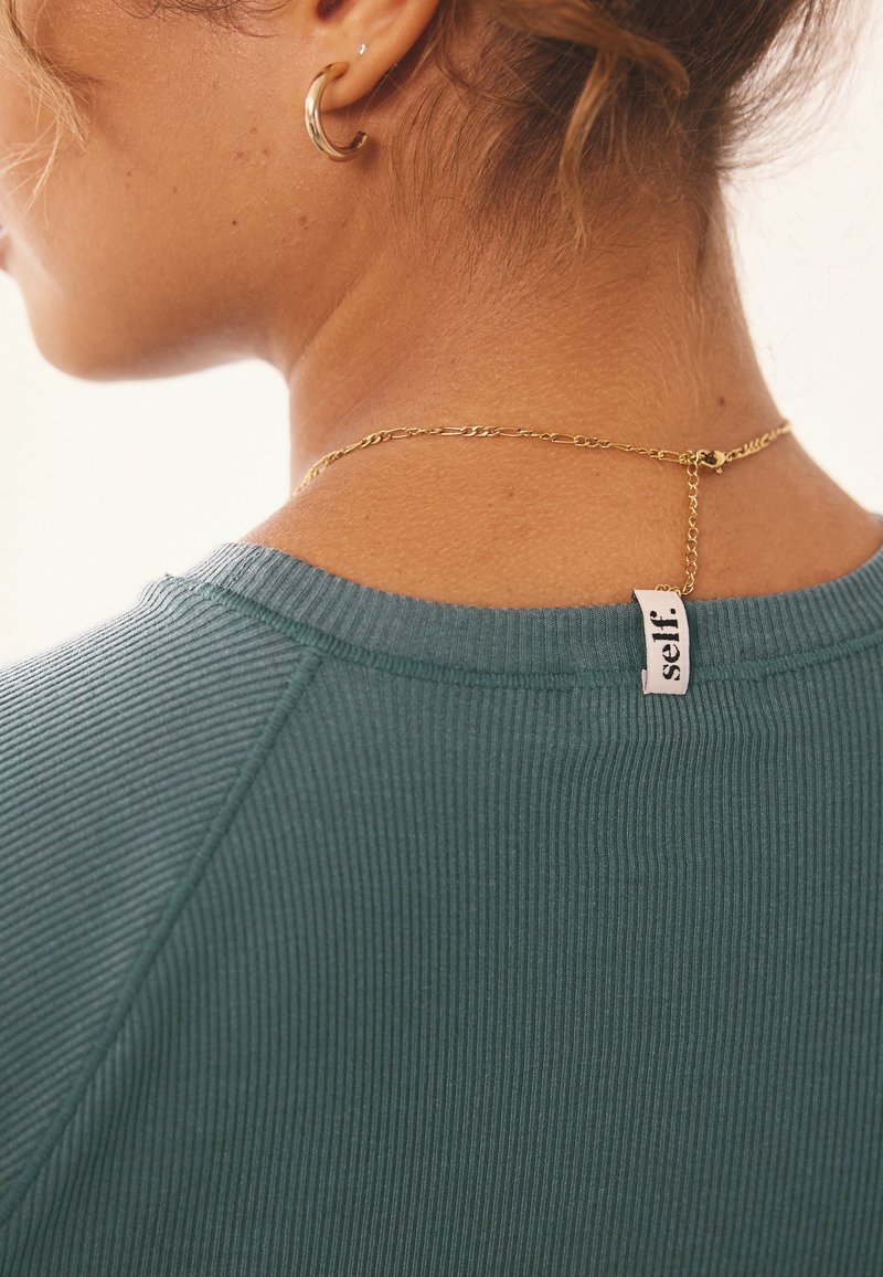 Femme aux cheveux blonds portant un haut côtelé vert, une boucle d'oreille en or en forme d'anneau et un collier en chaîne en or avec une étiquette "self." au col du haut.