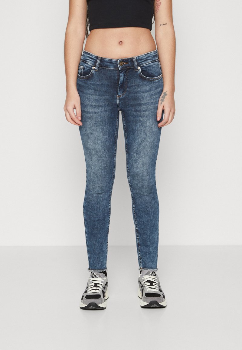 Jeans de denim azul ajustados con detalles de desgaste, diseño de cinco bolsillos y cierre de botón. Llevados con zapatillas deportivas en negro y gris.