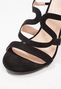 Sandale ouverte en daim noir avec des brides avant ondulées et une semelle intérieure beige rembourrée sur fond blanc.
