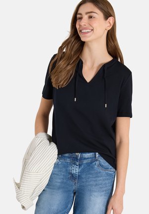 Femme souriante aux longs cheveux bruns portant un haut noir à manches courtes et un jean bleu, tenant une veste blanche rayée dans une main.