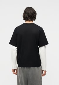 Άτομο με μακριά μέχρι τον ώμο μαλλιά που φορεί μαύρο πουκάμισο με κοντά μανίκια πάνω από λευκό πουκάμισο με μακριά μανίκια και γκρι παντελόνι, γυρισμένο με την πλάτη στην κάμερα.