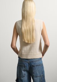 Person mit langen blonden Haaren, die ein ärmelloses beiges Strickoberteil und blaue Jeans trägt, steht mit den Händen in den hinteren Taschen.