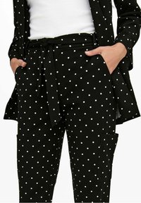 Black pants with white polka dots, a tied belt, and side pockets worn with matching polka dot jacket and white ribbed top. Hands in pockets.